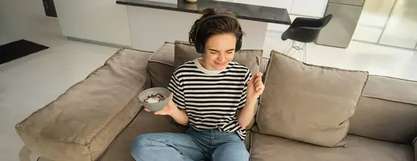 Portrait of carefree smiling woman, listens music in headphones, holding bowl with cereals, eating breakfast, sitting on sofa in living room and making dance moves.