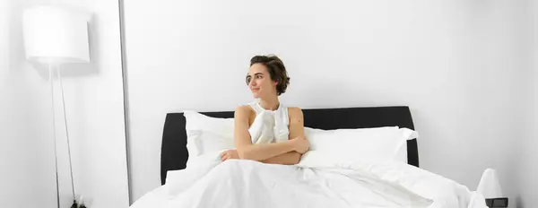 Portrait of young smiling woman with messy hair, sitting on her bed, covered in white linen sheets, looking aside with dreamy face expression, waking up in morning.