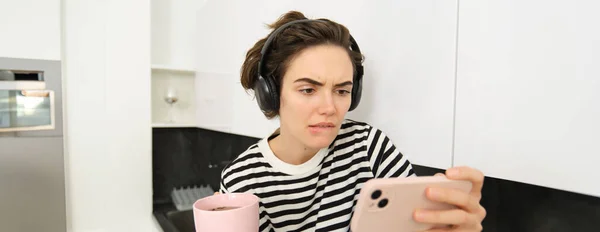 Portrait of woman with serious face, looking at smartphone with tensed emotion, watching interesting video on mobile phone, drinks tea, frowns and bites lips.