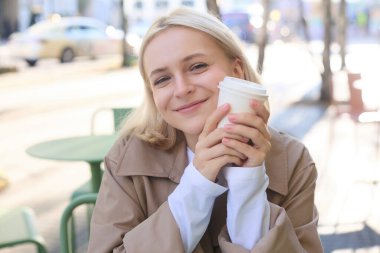 Gülümseyen sarışın kadının yakın portresi, elinde bir fincan kahve, açık havada, sokak kafesinde oturuyor, mutlu görünüyor, dışarıda restoranda birini bekliyor..