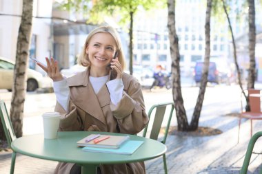 Havalı sarışın bir kadının cep telefonuyla konuşması, açık kafede kahve içmesi, şehir merkezinde sıcak havanın tadını çıkarması, akıllı telefondan telefona cevap vermesi..