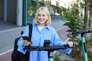 Genç gülümseyen bir kadının portresi, elektrikli scooter 'ın kilidini açıyor, akıllı telefon uygulaması ile QR kodunu tarayıp aracının kilidini açıyor, binanın yanındaki sokakta duruyor..