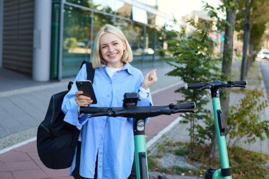 Güzel sarışın kız, cep telefonunda QR kodunu tarıyor, kiralamak için sokak scooter 'ının yanında duruyor, bir gezinti istiyor, gülümsüyor ve mutlu görünüyor..