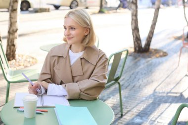 Modern bir genç kadın, proje üzerinde çalışan bir öğrenci, açık kafede oturuyor, kahve içiyor ve tek başına çalışıyor, ders çalışıyor ya da ödev yapıyor..
