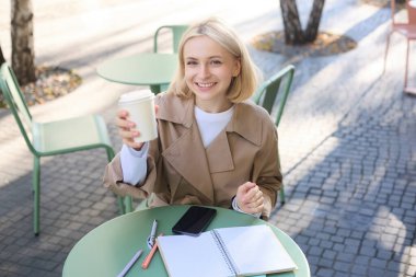 Güzel sarışın kadın, gülümsüyor, beyaz kahve fincanı gösteriyor, kafeteryada içki içiyor, dışarıda oturuyor..