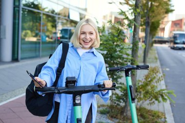 Genç gülümseyen bir kadının portresi, elektrikli scooter 'ın kilidini açıyor, akıllı telefon uygulaması ile QR kodunu tarayıp aracının kilidini açıyor, binanın yanındaki sokakta duruyor..
