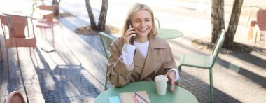 Neşeli genç bir kadın gülüyor, cep telefonuyla konuşurken gülümsüyor, açık kafede oturuyor, arkadaşça sohbetin tadını çıkarıyor..