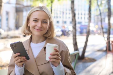 Akıllı telefonlu tatlı sarışın kadın portresi, çay içmek, şehir merkezindeki favori mekanda kahve, açık havada oturmak, cep telefonu tutmak..