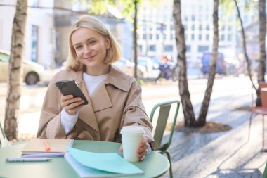 Akıllı telefonlu, gülümseyen sarışın bir kadının portresi, elinde bir fincan kahve, çay içmek ve şehir merkezinde güneşli bir günün tadını çıkarmak..