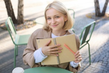 Defterli güzel sarışın bir kızın açık havada çekilmiş fotoğrafı, açık kafede oturan bir kadın, günlüğe yazıyor, ajandasına planlar yapıyor, notlar yazıyor..