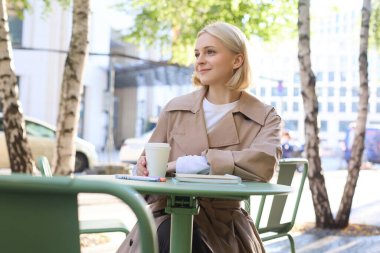 Şehir yaşam tarzı konsepti. Genç ve güzel bir kadın açık hava kafede oturuyor, şehir hayatını düşünüyor, kahve içiyor ve sokakta gülümsüyor..