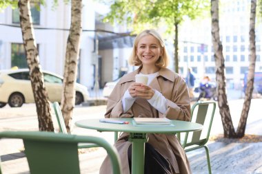 Modern bir kadın portresi, açık bir kafede oturuyor, kahve içiyor ve gülümsüyor, trençkot giyiyor..