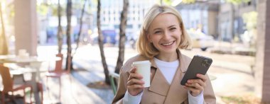 Akıllı telefonlu, gülümseyen sarışın bir kadının portresi, elinde bir fincan kahve, çay içmek ve şehir merkezinde güneşli bir günün tadını çıkarmak..