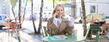 Havalı sarışın bir kadının cep telefonuyla konuşması, açık kafede kahve içmesi, şehir merkezinde sıcak havanın tadını çıkarması, akıllı telefondan telefona cevap vermesi..