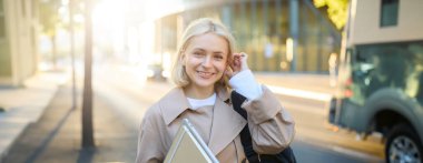 Sarışın genç bir kadın, kolej öğrencisi, elinde günlükler ve defterler, ödevlerini taşıyor, üniversiteye gidiyor, gülümsüyor ve mutlu görünüyor..