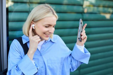 Sarışın, gülümseyen kadın modelin yakın portresi, kulaklıklı üniversite öğrencisi, sokakta müzik dinleyen, akıllı telefon tutan, dışarıda birini bekleyen....