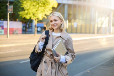 Modern bir kadının resmi, sırt çantası ve defterleri olan bir öğrenci, kadın model, işe gidiyor, güneşli bir sonbahar gününde şık bir siper giyiyor..