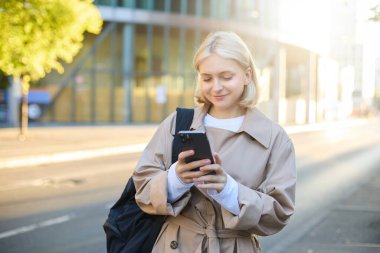 Sarışın, gülümseyen bir kadının sokak tarzı portresi sokakta yürür, güneşli, aydınlık bir gün, elinde akıllı bir telefon, sırt çantası taşır, cep telefonuyla taksi çağırır, cep telefonundan mesaj gönderir..