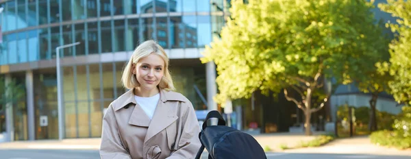 Vertical shot of young female employee, woman sitting on street bench with backpack and smartphone, waiting for her taxi, using mobile app, looking confident at camera.