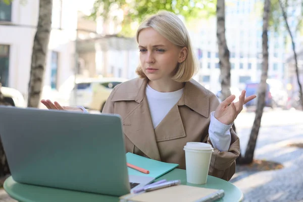 Image of woman with confused face, looking at laptop, working remotely, chatting with someone and shrugging, talking with puzzled face, sitting in coffee shop.