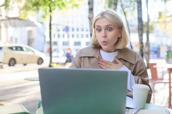 Close up portrait of young blond woman, sitting outdoors with laptop, drinking coffee in cafe, looking amazed and surprised at device screen, hears awesome news.