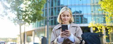 Genç sarışın kadın portresi, sokak bankında oturan, akıllı telefonlu, cep telefonu uygulamalı birini bekliyor..