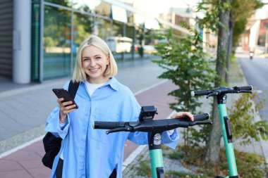 Genç bir kadının portresi, scooter kiralamak, kilidi açmak için cep telefonu uygulaması kullanmak, işe gitmek için hızlı bir sürüş kullanmak, gülümsemek ve mutlu görünmek..
