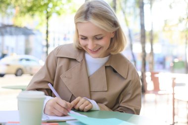 Modern bir genç kadın, proje üzerinde çalışan bir öğrenci, açık kafede oturuyor, kahve içiyor ve tek başına çalışıyor, ders çalışıyor ya da ödev yapıyor..