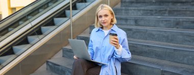 Merdivenlerde oturan genç bir kadının portresi bir fincan kahve ve dizüstü bilgisayarla çalışıyor, açık havada çalışıyor, serbest çalışıyor..