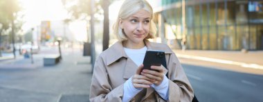 Sokakta duran genç sarışın kadının portresi, kararsız, şüpheli bir yüz ifadesi var, cep telefonu kullanıyor, isteksizlik gösteriyor..