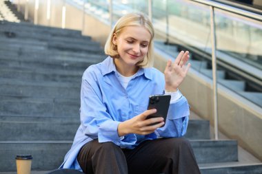 Gülümseyen sarışın kadının portresi, merdivenlerde bir fincan kahveyle oturuyor, akıllı telefona el sallıyor, görüntülü sohbete bağlanıyor, cep telefonuyla konuşuyor..