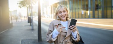 Şehrin sokaklarında genç bir kadının portresi, elinde akıllı telefon, cep telefonunu işaret ediyor, mutlu görünüyor ve kameraya gülümsüyor..