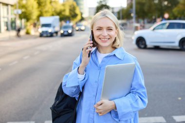 Mavi gömlekli, modern sarışın bir kadının resmi. Laptop ve sırt çantası var. Akıllı telefon kullanıyor, gülümsüyor, cadde boyunca yürüyor, şehir merkezinde poz veriyor..