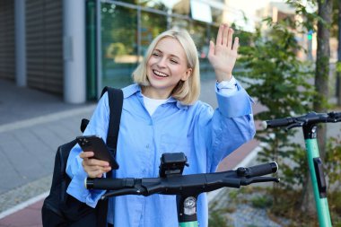 Genç, neşeli bir kadın, sokakta elektrikli scooter 'da QR kodunu taramaya çalışmış, onu kiralamak için cep telefonunu kullanmış, arkadaşına merhaba demiş ve birine el sallamış..