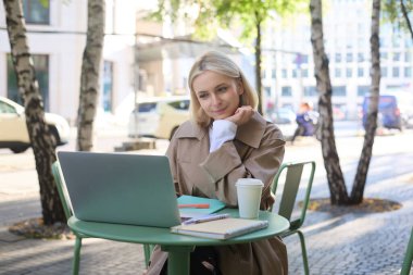 Dizüstü bilgisayara bakan, açık kafede oturan, kahve içen, uzaktan eğitim gören, dışarıda oturmuş, serbest çalışan genç bir kadının görüntüsü..