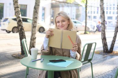 Modern bir kadının resmi, kafeteryada oturan bir öğrenci, elinde not defteri, sarı günlüğünü gösteriyor, planlayıcıyla yazıyor ve gülümsüyor..