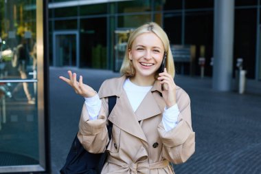 Akıllı telefonlu kaygısız genç bir kadın, şehir merkezinde dışarıda yürüyor, caddede duruyor ve sohbet ediyor, cep telefonuyla konuşuyor, mutlu, güler yüzlü bir yüz ifadesiyle..