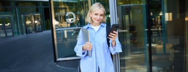 Üniversite kampüsünün önünde duran genç kız öğrencinin portresi, binanın yanında poz veriyor, dizüstü bilgisayar ve akıllı telefon tutuyor, mavi yakalı gömlek giyiyor..