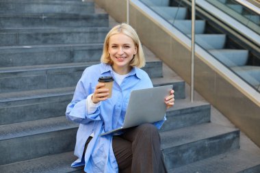 Genç bir kadın elinde bir fincan kahve ve dizüstü bilgisayarla dışarıda merdivenlerde oturuyor, uzaktan çalışıyor, serbest çalışıyor, internetten çalışmak için şehirdeki internet bağlantılarına bağlanıyor..