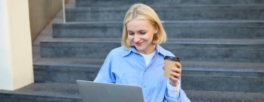 Gülümseyen genç şehir kadını, dizüstü bilgisayarda çalışan öğrenci, merdivenlerde oturuyor, kahve içiyor, çalışıyor, temiz havada e-öğreniyor..