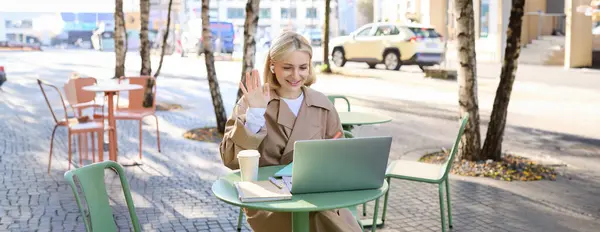 Cheerful girl with laptop, saying hello, connects to online meeting, waving hand at camera, chatting with someone via internet, sitting in outdoor coffee shop.