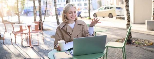 Friendly, smiling blond woman chatting online, wearing wireless headphones, looking at laptop and gesturing, talking to someone, working remotely.