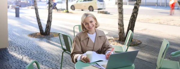 Stock image Image of young blond modern woman, sitting with laptop outside in cafe, drinking coffee drink and working on project, using computer.