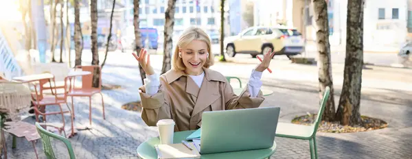 Enthusiastic young woman hear great news during online meeting, looking at laptop and celebrating, raising hands up to cheer, sitting in outdoor cafe.