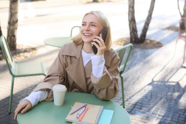 Neşeli genç bir kadın gülüyor, cep telefonuyla konuşurken gülümsüyor, açık kafede oturuyor, arkadaşça sohbetin tadını çıkarıyor..