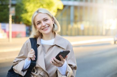 Sırt çantalı, akıllı telefonlu, kameraya mutlu bir şekilde bakan, sokakta bekleyen, sırtüstü gülümseyen üniversiteli genç bir kızın yakın portresi..