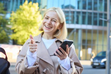 Neşeli genç sarışın kadın, üniversite öğrencisi, bankta oturuyor, kameraya başparmağını kaldırıyor ve göz kırpıyor, cep telefonu tutuyor, güzel bir şey öneriyor..