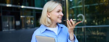 Gülümseyen sarışın güzel bir kız, dizüstü bilgisayarı olan bir ofis çalışanı akıllı telefon uygulamasından sesli mesaj kaydediyor, şirket binasının yanında duruyor..