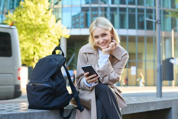Lifestyle portrait of smiling female model sitting on street bench with backpack and mobile phone, waiting for friend and scrolling social media on smartphone.