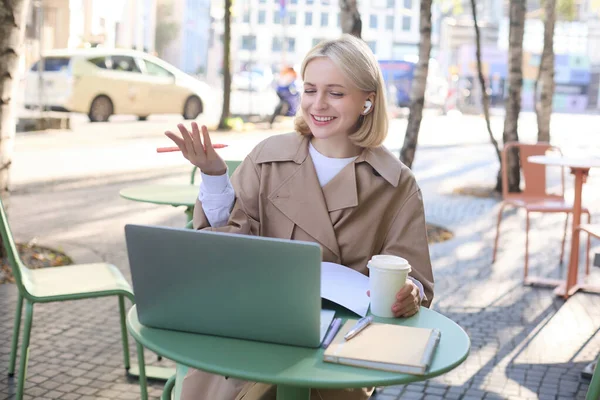 Friendly, smiling blond woman chatting online, wearing wireless headphones, looking at laptop and gesturing, talking to someone, working remotely.